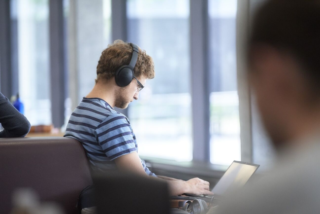 Ein junger Mann in T-Shirt und einem Kopfhörer auf dem Kopf arbeitet auf seinem Laptop-Computer in der Lounge des PHBern-Gebäudes
