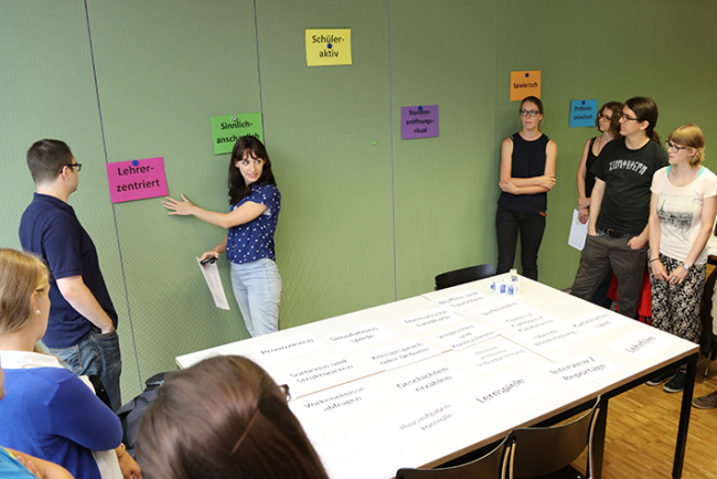 Studierende des Instituts Sekundarstufe II im Seminar Allgemeine Didaktik. Eine Studentin steht an einer Magnetwand, an der Zettel mit Überschriften wie „Lehrerzentriert“ hängen und erklärt etwas.